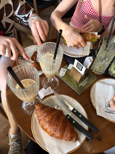 In Ulcinj, Montenegro, a group of people are enjoying brunch at a table. The image focuses on a wooden table laden with food and drinks. In the foreground, a large croissant sits on a white plate with a brown rim, accompanied by a knife and fork. To the left, a glass filled with iced coffee and a straw is prominently featured. Another glass, also containing iced coffee and a straw, is visible in the mid-ground, along with a partially eaten croissant on a plate.
Further back, a person in a red and white checkered top is holding a croissant sandwich, suggesting they are either preparing to eat it or have just taken a bite. Another individual, wearing a brown and white patterned shirt, is using a knife and fork to cut into food on a plate. The table also holds glasses of drinks, cutlery, napkins, and a menu with a QR code. The environment appears to be an outdoor cafe setting, with hints of a sunny day. The overall scene captures a relaxed brunch gathering.