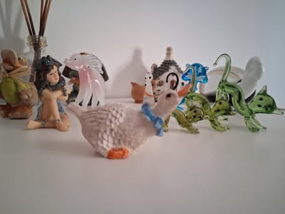 The image shows a collection of decorative objects hanging from a white ceiling, possibly in a home or a shop in Colorno, Italy. The objects are diverse and appear to be made of glass, ceramic, and other materials.
On the left, there are several green glass objects that resemble leaves or abstract shapes, some with dark spots. Further to the right, there is a white object that could be a stylized bird or a cloud. In the center, a large, textured object that looks like a white bird with an orange beak or cap dominates the scene. Below this bird, there's a smaller, more detailed ceramic figure that appears to be a house with a chimney and a small character with black dots for eyes hanging from it.
To the right of the central bird, there are more whimsical figures. A translucent pink glass bird with detailed wings is prominent. Next to it, a figurine of a woman with dark hair, wearing a blue necklace and a tan skirt, is suspended upside down. To her right, another upside-down figurine appears to be a duck or a similar bird, wearing a green garment and tied with a straw ribbon. These figures are hanging from a string or wire attached to a glass bottle at the bottom right, which has some wooden sticks emerging from it.
The lighting suggests it might be daytime, with the light source coming from above, casting subtle shadows on the white surface. The overall impression is one of a quirky and eclectic collection of decorative items. No text is visible in the image.