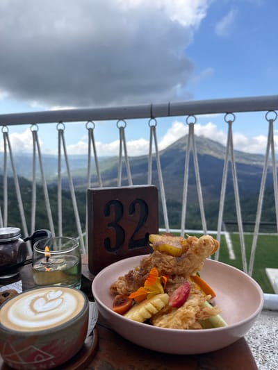 This is a close-up shot of a table at a restaurant in Kintamani, Indonesia, with a scenic mountain view. The table is set with a coffee cup featuring latte art, a small candle in a glass of oil, a jar of coffee grounds, a wooden table number that reads "32," and a pink bowl filled with food. The food appears to be a fried item, possibly chicken or fish, accompanied by sliced fruits like pineapple, strawberry, and orange, and some green vegetables.
In the background, a railing is visible, beyond which lies a breathtaking landscape. Rolling hills covered in lush greenery stretch into the distance, culminating in a majestic mountain, likely Mount Batur, dominating the horizon under a cloudy sky. The overall impression is one of a relaxed dining experience with a stunning natural backdrop, typical of Kintamani's popular viewpoints.