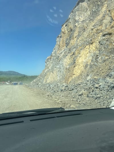The image depicts a construction site with a large rock face on the right. The ground is unpaved and dusty, with a dirt road leading into the distance on the left. There are some portable structures and vehicles visible on the left side of the road. In the background, there are rolling hills covered in greenery, with a clear blue sky above. The image appears to have been taken from inside a vehicle, as the dashboard is visible in the foreground. There are no people clearly visible in the image. The location is identified as Sarasa, Russia.