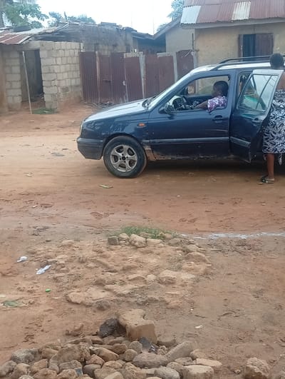 The image depicts a daytime scene in Yelwa, Nigeria, focusing on a blue four-door car parked on a dusty, unpaved road. A person, likely a woman, is seated in the driver's seat, wearing a light-colored, patterned top, and appears to be looking forward with her hands on the steering wheel. The rear passenger door on the same side is open, and another person, also likely a woman, is partially visible standing next to the car, wearing a patterned skirt or dress and dark flip-flops.
In the background, several buildings contribute to the scene's character. To the left, there's a structure made of unrendered concrete blocks, possibly unfinished or under construction, with an open doorway. Adjacent to it are sections of corrugated metal, appearing as fencing or gates. Further back and to the right, another building with a light-colored, plastered wall and a corrugated metal roof is visible. Sparse green foliage is seen above these structures under a bright, possibly overcast sky.
The ground is a prominent feature, consisting of reddish-brown dirt, uneven and scattered with numerous rocks, including a large pile in the immediate foreground. Small pieces of litter, such as white plastic items and a green cap, are also visible on the ground. The car itself shows visible dirt and dust, consistent with travel on unpaved roads. The overall image captures a candid moment, suggesting a brief stop or activity involving the car and its occupants in a semi-urban or rural setting. No discernible text is visible in the image.