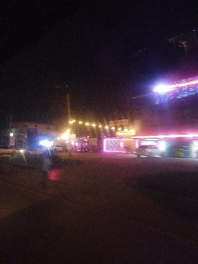 A dimly lit street scene at night reveals various illuminated buildings with bright storefronts and signs. Several buildings display a series of windows with warm, yellow lights emanating from within. A Ferris wheel-like structure is outlined by colorful lights. The ground is dark and undefined, adding to the overall low-light atmosphere.