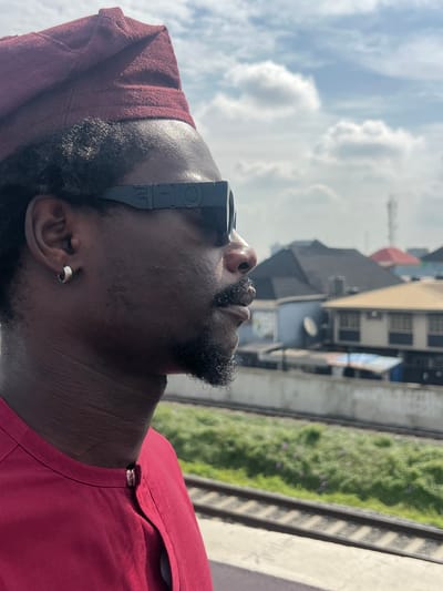 This vertically oriented image captures the profile of an adult male looking upwards towards a cloudy sky. The subject has dark skin, a neatly trimmed beard, and is wearing a maroon-colored shirt with a matching head covering or cap. A small silver hoop earring is visible in his left ear. He is wearing a unique pair of black glasses or goggles, with the text "IOE" visible on the side temple.
The background, which is out of focus, reveals an urban or suburban setting. Several buildings are visible, including one with multiple windows and another with a dark roof. Below these structures, what appear to be train tracks or a raised concrete platform with green vegetation run parallel across the mid-ground. The sky above is bright with scattered white cumulus clouds, indicating daytime. The man's upward gaze suggests he is observing something in the sky or distance. The overall scene is consistent with an outdoor location in Ikeja, Nigeria.