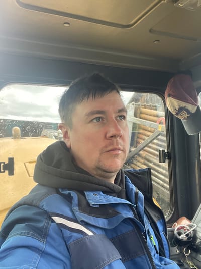 A man is pictured inside the cab of a piece of heavy machinery, potentially a tractor or excavator, as suggested by the yellow equipment visible through the window. He is wearing a blue work jacket over a dark hooded sweatshirt. Above him, affixed to the cab, is a baseball cap and what appears to be a small, possibly decorative, object resembling a soda can. The view through the window shows an exterior scene with stacked logs. The location of the picture is the city of Поедуги, Russia.
