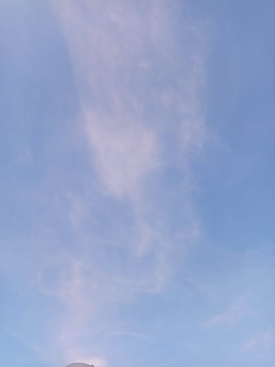 This is a vertical, eye-level, medium shot photograph of a pale blue sky with wispy, light pink and white clouds. The clouds are thin and spread out across the sky, with a more concentrated patch of cloud formation in the upper center of the frame. The bottom edge of the image shows the top of a white, curved structure, possibly the roof of a building or a dome, partially visible against the sky. The scene is devoid of any human presence or activity. The lighting suggests it might be dawn or dusk, given the soft, pastel hues in the sky. The location is Pampatar, Venezuela.