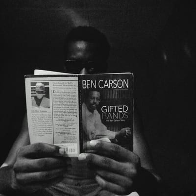 A person is reading the book "Gifted Hands" by Ben Carson. The book is open to the front cover and an inside page that contains a photo of Dr. Ben Carson and text that includes the blurb "From Inner-City Kid to Renowned Neurosurgeon." The person's face is partially visible above the book, appearing to wear glasses. The scene is black and white and grainy. There is no explicit reference to the location being Keffi, Nigeria.