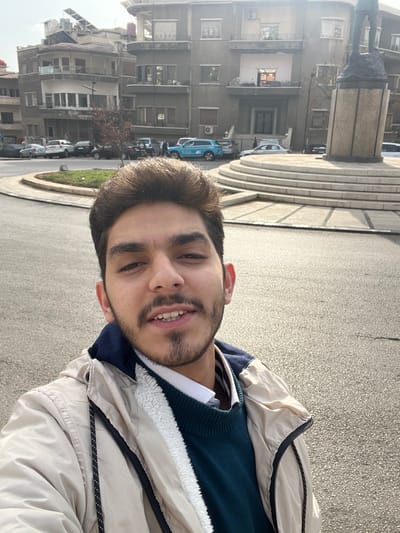 A young man takes a selfie outdoors in what appears to be Al-Muhajerin Municipality, Syria, during a bright daytime. He looks at the camera with a slight smile, having dark hair and a neatly trimmed beard. He is dressed in a beige jacket featuring a furry white lining, worn over a dark blue sweater and a white collared shirt.
The background reveals an urban setting. To the left and behind the man, several multi-story buildings with windows and balconies are visible, characteristic of residential or mixed-use architecture, with some air conditioning units attached to their facades. In the mid-ground, a prominent, round, tiered stone structure, possibly a monument base, stands with a dark-colored statue or sculpture on top. A paved street or plaza occupies the foreground and mid-ground, where several cars are parked, including a distinct light blue car. Patches of green landscaping are also present. The scene suggests a casual moment on a clear day in a bustling city environment. No discernible text is visible.