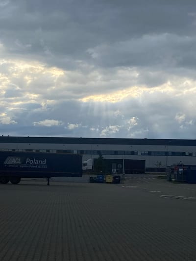The image depicts a truck trailer with "ACL Poland" printed on its side, parked in front of a long industrial building with multiple windows. Above the building, the sky is filled with dramatic clouds, with shafts of sunlight breaking through, creating a crepuscular ray effect. The ground in the foreground is paved with dark interlocking stones, suggesting an outdoor area like a loading dock or parking lot. In the distance, there are some recycling bins and traffic signs, including a stop sign. The overall impression is one of a logistics or distribution center under a moody sky. The location is identified as Bielany Wrocławskie, Poland.