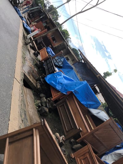 The image shows a street side in Arusha, Tanzania, at coordinates -3.363, 36.678. It appears to be a furniture store or workshop, with several wooden bed frames stacked on top of each other and covered with blue tarpaulin. The frames are in various stages of construction, and some are leaning against a building. The scene is cluttered, with some pieces of wood and debris scattered on the ground. There are some trees in the background, and the sky is partly cloudy. The image was taken from a low angle, looking up at the bed frames. The bed frames are all made of dark wood and appear to be new. There are no people visible in the image.