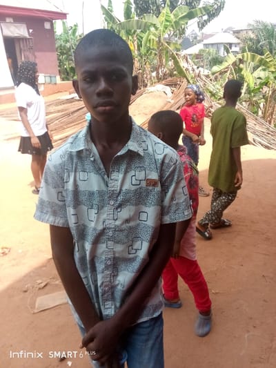 The image portrays an outdoor scene within Akpaka Forest Reser, Nigeria, featuring five individuals and an unpaved environment. In the foreground, a young male, approximately 12-15 years old, with dark skin and a short buzz cut, stands centrally, directly facing the camera. He wears a light-colored, short-sleeved button-up shirt with dark grey abstract patterns of wavy lines and squares, over blue jeans. His hands are clasped in front of him, and his expression is neutral.
To the left and slightly behind the foreground subject, a female with dark skin, wearing a white short-sleeved top and a dark skirt, is visible. Her hair is covered by a dark, patterned headscarf, and she stands facing left. Behind her, a light-colored building with a dark roof and a window is partially visible.
In the mid-ground, to the right of the central figure, several other children are present. One child, wearing a red t-shirt and blue jeans with a patterned head covering, faces right with an open mouth, as if speaking or shouting. Another child, largely obscured by the foreground subject, wears a patterned red top and red pants. A third child, wearing a green t-shirt and patterned green pants with sandals, walks away from the camera towards the right.
A large pile of reinforcing bars (rebar) is situated in the mid-ground behind the children. The background consists of dense green vegetation, including large-leafed plants resembling banana trees, with a few distant residential buildings featuring light-colored roofs. The ground is reddish-brown, unpaved earth. The scene is brightly illuminated by natural daylight.