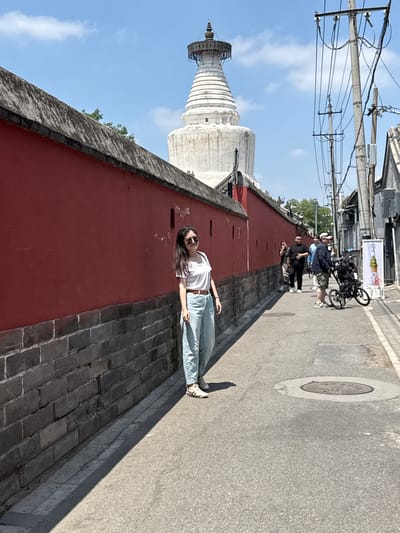 In the Xicheng District of China, a young woman stands on a paved alleyway, leaning against a weathered red brick wall. She wears a white t-shirt, light blue jeans, and sunglasses. The alley is flanked by a high, red-painted wall on the left and buildings on the right, interspersed with utility poles and wires. In the distance, a prominent white stupa with tiered levels rises against a bright blue sky with scattered clouds. Several other people are visible further down the alley, some walking and one with a stroller. A sign displaying an image of green soft-serve ice cream stands near the right side of the alley. The ground is asphalt with a circular manhole cover. The overall scene suggests a sunny day in an urban environment with historical architecture.