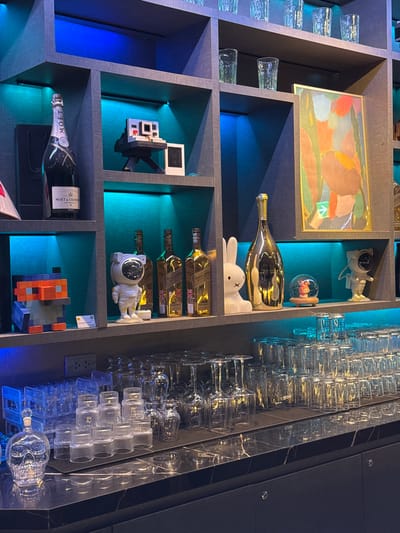 The image depicts a dimly lit bar or lounge area. On a shelf, a bottle of Moët & Chandon Imperial Brut champagne stands next to a black box and a Polaroid camera replica. Another shelf displays three bottles of Johnnie Walker Gold Label Reserve whisky, a white bunny figurine, a golden bottle, a snow globe with a figurine inside, and a small astronaut figurine. Further down, on the counter, is a collection of drinking glasses, some stacked and others inverted, and a glass decanter shaped like a skull. The shelves are illuminated with blue and teal LED lighting. The overall impression is of a sophisticated establishment, possibly a bar in Hanoi, Vietnam, given the context provided.