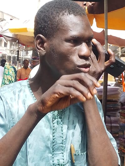 The image depicts an outdoor market scene in Fagge A, Nigeria, during daylight hours. A Black male, appearing to be in his 20s or 30s, is positioned in the foreground, facing right in profile, with his head turned slightly towards the viewer. He wears a light blue traditional garment with embossed patterns and decorative stitching around the collar and chest. A light-colored object, resembling a pen, is clipped into his left breast pocket. He holds a black mobile phone to his right ear with his right hand and has his left hand raised, fingers curled, near his mouth. His mouth is slightly agape. In the blurred background, market activity is visible, including several individuals, one wearing a green and white patterned garment, another a light brown traditional outfit. Multiple large, open market umbrellas, predominantly yellow and orange, provide shade. Stacks of folded fabrics are visible on the right, and multi-story buildings line the distant background.