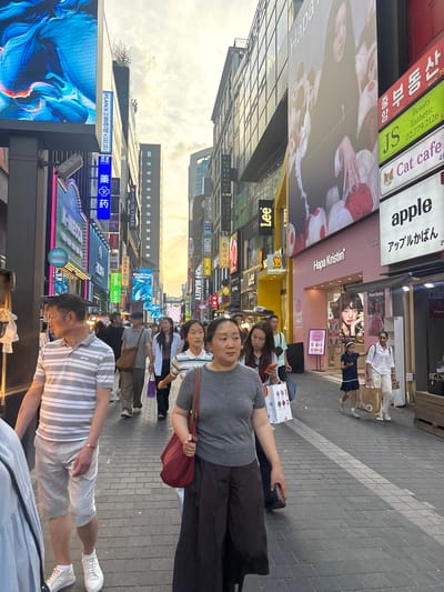 The image depicts a bustling street scene in Myeongdong, Seoul, South Korea, during daylight hours. A diverse group of people are walking along a paved pedestrian walkway lined with shops and large digital billboards. The architecture is modern, with multi-story buildings showcasing bright signage in Korean and English.
Several individuals are the primary focus. In the foreground, a woman with dark hair is walking towards the viewer, carrying a large red tote bag. To her left, a man in a grey and white striped polo shirt and checkered shorts is also walking. Behind them, other pedestrians, including women and children, are seen moving through the crowd, some carrying shopping bags.
The street is filled with commercial activity. Shops display vibrant advertisements and illuminated signs. Prominent brands and store names like "Hapa Kristin" and "apple" are visible on the buildings. The overall atmosphere suggests a popular shopping district, with a constant flow of people engaged in leisurely activities and commerce. The sky above is partly cloudy, indicating a mild weather day.
