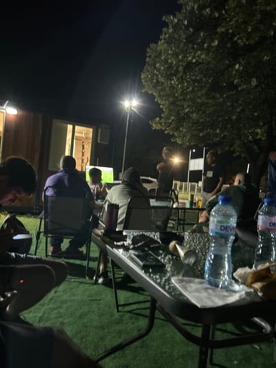 An outdoor gathering is taking place at night, possibly in Vratsa, Bulgaria, indicated by the presence of people watching a television screen displaying what appears to be a sports match. The scene includes a portable building with windows, outdoor lighting illuminating the area, and a large tree with foliage. Several individuals are seated around a glass-top table, with some drinks and other items visible on the table. A green artificial turf surface is present. One person in the foreground is looking at their phone. The overall atmosphere suggests a relaxed, casual social event.