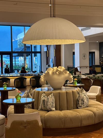 The image depicts a luxurious interior space, likely a high-end hotel lobby or lounge. Features include a large, off-white, dome-shaped lamp fixture dominating the left foreground, partially obscuring the background. To the right is a large, curving sofa with patterned pillows. Glass-topped tables with floral arrangements are arranged near the seating. Through large windows, a partial view of a city environment with a possible outdoor seating area is visible. The architecture suggests a contemporary design aesthetic. People are present in the scene, engaged in activities consistent with a lobby environment. The location is identified as Cape Town, South Africa based on the architecture.