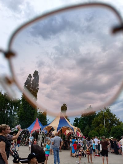 The image depicts an outdoor gathering, possibly a festival or market, in Orenburg, Russia. The foreground is dominated by the blurred rim of eyeglasses, through which the scene is viewed. Behind the glasses, a crowd of people are visible, interacting and moving around. Several colorful tents, likely stalls or performance areas, are set up in the background. Trees and a cloudy sky complete the environment. The atmosphere appears casual and social, with individuals of various ages present.