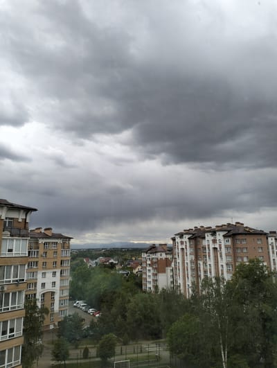 The image shows an urban scene under a dark, overcast sky, likely in Krykhivtsi, Ukraine. Two multi-story residential buildings dominate the foreground, one on the left and one on the right, constructed in a similar architectural style, with brick and white facade elements. Between the buildings, there are trees and a view of distant houses and hills. Parked cars are visible in a designated parking area amongst the trees. The sky is filled with heavy, dark clouds, indicating possible rain. A faint silhouette of mountains is visible in the background. A playground is at the very bottom with a fence.