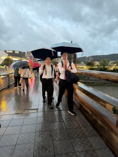 The image captures an outdoor urban scene at dusk or night during rainfall, specifically on the Sanjo Ohashi Bridge over the Kamo River in Kyoto, Japan. The sky is uniformly grey and overcast. The bridge surface is visibly wet and reflective.

In the immediate foreground, two East Asian males are walking towards the viewer from the middle-ground. The male on the viewer's right is partially blurred due to motion. He wears a white button-down shirt, dark trousers, and a purple tie, carrying a black shoulder bag and holding a black umbrella over his head. The male to his left, slightly further back and in sharper focus, wears a white button-down shirt, dark trousers, and a dark backpack, also holding a black umbrella.

Further into the background on the bridge, additional pedestrians are visible, moving in various directions. They are also carrying open umbrellas, some black, one distinct red with a pattern, and others of lighter hues. The bridge features wooden railings on both sides, which appear wet and reflective. Below the bridge, water from the Kamo River is discernible.

On the left side of the frame, beyond the bridge, city buildings are present, displaying illuminated signs. One prominent sign clearly displays the Japanese characters '三条駅' (Sanjo Station). A red traffic light is also visible. In the background, dark, tree-covered hills rise under the low, cloudy sky, framing the cityscape.