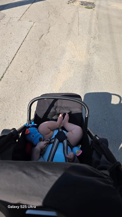 The image shows a baby lying in a black stroller. The baby is wearing a blue outfit and is secured with a harness. A blue toy is attached to the stroller. The stroller is on a paved surface, likely a sidewalk or path, with shadows cast by an unseen object, suggesting it is daytime. The weather appears sunny. In the background, the ground is visible, with some cracks and a manhole cover. The watermark "Galaxy S25 Ultra" is present on the right side of the image. The scene is outdoors, possibly in Ezerovo, Bulgaria, as per the provided location context. There is no indication of a specific event taking place, other than the baby being taken for a walk or outing. The overall mood appears peaceful.