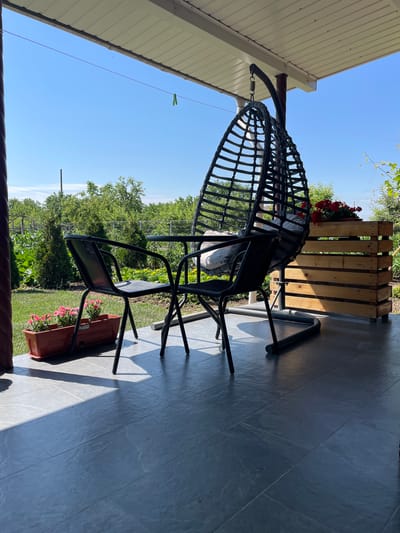 The image shows a porch area with outdoor furniture. A black wicker hanging egg chair is suspended from a metal stand. In front of it are two black metal chairs and a small table. To the left, a rectangular brown planter holds pink flowers. To the right, a wooden planter box contains red flowers. The porch floor is tiled with large, grey tiles. Beyond the porch, a green lawn is visible, with various trees and plants in the background. The sky is clear and blue. The scene appears to be a residential outdoor space.