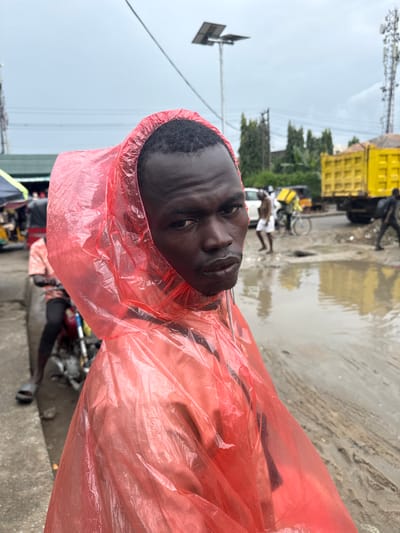 A medium shot captures a man wearing a red plastic poncho, his head and shoulders dominating the foreground. He is looking directly at the camera with a neutral expression. The poncho is translucent, revealing hints of the clothing underneath and the texture of the plastic. The background depicts a street scene in Lagos, Nigeria, characterized by wet, muddy ground and large puddles reflecting the overcast sky and surrounding elements. In the mid-ground, a yellow truck is visible, along with other vehicles and a few people. The overall atmosphere suggests it is a rainy day in a busy urban environment.