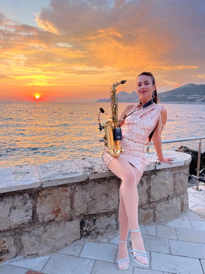 A woman sits on a stone wall overlooking the Adriatic Sea at sunset. She holds a saxophone, and her dress is covered in sequins. The sky is filled with orange and yellow clouds, and the sun is setting over the water. In the background, mountains rise from the sea. The location is identified as Pečurice, Montenegro.