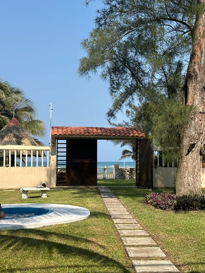A daytime shot of a landscaped beachfront property in Playa de Chachalacas, Mexico. A tan-colored building with a thatched roof and white railings is visible in the foreground. A dark, square structure with a red tiled roof stands on a grassy area next to a stone-paved path. The ocean and beach are visible through the structures. A large tree is on the left. A white surfboard lies on the grass.