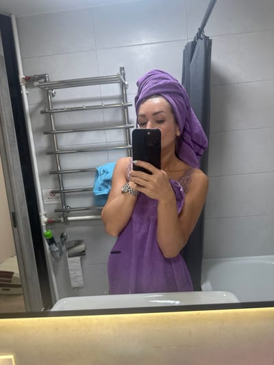 A woman in a bathroom, wrapped in a purple towel, takes a selfie in a mirror. She has a purple towel wrapped around her head. She is wearing a light-colored lace top under the towel. She holds a black smartphone in her hands, taking the picture. The bathroom has tiled walls, a metal towel rack with multiple bars, and a gray shower curtain. A white bathtub is partially visible. In the foreground, the edge of a shelf or counter is illuminated by a light fixture. The scene suggests a personal moment of self-care or preparation after showering.