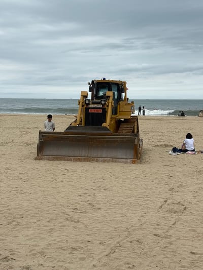 The image depicts a beach scene with a large yellow bulldozer in the foreground. The bulldozer is positioned in the sand, facing the ocean. Behind the bulldozer, the sea is visible with waves rolling in under a cloudy sky. Several people are present on the beach. A young man in a light-colored t-shirt stands to the left of the bulldozer, looking down. To the right, a person in a white top is sitting on the sand with their legs crossed, facing away from the camera. In the distance, other figures can be seen near the water's edge. The sand is light brown and shows some footprints and textures. The location appears to be a public beach, likely in Busan, South Korea, given the context.