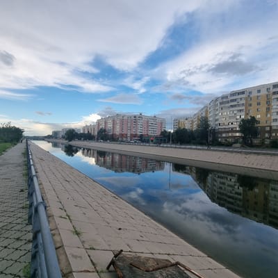 The image shows a riverside scene in Aktobe, Kazakhstan. The camera is tilted, giving the impression that the water and the sky are a single, reflective surface. On the left, tall apartment buildings with many windows are visible, reflecting in the water. The buildings vary in color, with some appearing yellow and others pink. Trees are interspersed between the buildings. On the right side of the image, a paved walkway runs alongside the water, made of large, square concrete tiles. A metal railing runs along the edge of the walkway, and further to the right, a piece of rusted metal, possibly part of a bench or a discarded object, is visible. The sky is partly cloudy, with patches of blue sky visible between white and gray clouds. The reflection of the sky and buildings in the water is quite clear, suggesting calm water. The overall lighting suggests it might be late afternoon or early evening. There are no visible people in the image, but the presence of a walkway and railing indicates this is a public area. The scene evokes a sense of urban tranquility, with the natural element of the water reflecting the man-made structures and the sky.