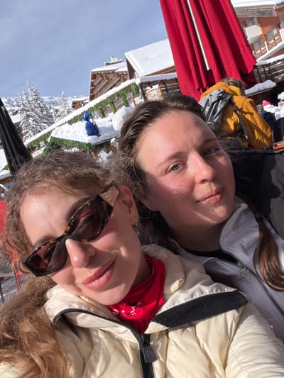 Two females are photographed outdoors in a snowy alpine environment, specifically in the city of Les Allues, France. The foreground features the upper torsos and faces of two individuals.
The person on the left has light brown, curly hair and wears dark tortoise-shell cat-eye sunglasses with gold accents on the temples. She is dressed in a light-colored puffer jacket, possibly cream or off-white, with a red bandana visible around her neck. Her lips are slightly parted in a subtle smile.
The person on the right has dark brown hair, pulled back, and discernible blue eyes. She wears a light grey or white ski jacket over a dark layer and looks directly towards the camera with a neutral expression.
The background consists of multiple snow-covered wooden chalet-style buildings, characteristic of a ski resort. These buildings have snow-laden roofs and balconies decorated with green garlands and string lights. A blue decorative object rests on a snow-covered ledge of one building. Snow-covered evergreen trees are visible on the mountain slopes behind the structures. Portions of large red and black outdoor umbrellas are also present. The sky is clear blue, indicating a sunny day. Distant figures in winter clothing are visible further in the background, suggesting an active outdoor area.