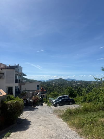 A daytime, medium shot captures a view overlooking a lush, green hillside dotted with buildings, leading to a distant hazy ocean and mountains under a clear blue sky with wispy clouds. In the foreground, a paved road curves downhill from a modern, multi-story building with balconies and spiral staircases on the left. Two cars, a dark sedan and a dark SUV, are parked on the right side of the road, partially obscured by overgrown green foliage. The building appears to be situated in a residential or resort area, possibly on a slope, with a driveway leading up to it. The overall scene suggests a tropical environment, consistent with the location of Baan Chaweng, Thailand.