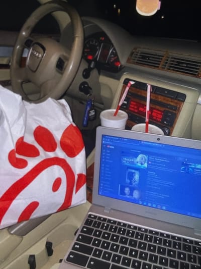 This image depicts the interior of a vehicle, likely an Audi based on the steering wheel emblem, at night. The scene is set up for entertainment and dining, with a laptop open on the center console displaying what appears to be a YouTube video with movie trailers. In front of the laptop are two cups with straws, suggesting beverages. To the left, a white paper bag with the red Chick-fil-A logo is prominently featured, indicating a meal from that establishment. The car's dashboard and air vents are visible, along with the steering wheel and part of the driver's side door. Keys are hanging from the ignition. There is no indication of the specific location within Iquique, Chile, other than the general context of being inside a car. No people are visible in the image.