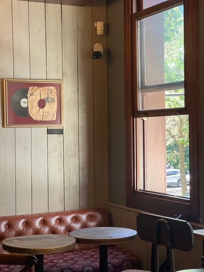 The image depicts an interior scene, likely a cafe or restaurant, with a framed vinyl record on a wooden paneled wall. A tufted brown leather sofa is visible in the foreground, along with two wooden tables. To the right, a large window reveals an outdoor view of trees and a car parked on a street. Two security cameras and a white device are mounted on the wall above the framed record. The lighting suggests it is daytime. The location is specified as Beşiktaş, Turkey.