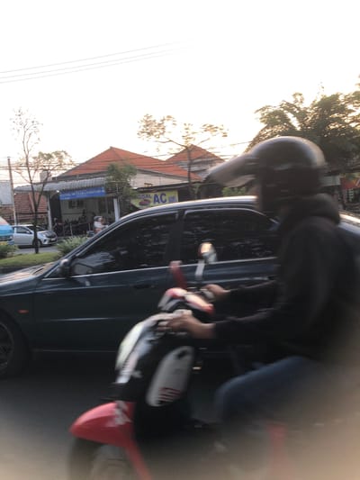 An outdoor, eye-level, medium shot captures a motorcyclist in motion, their face obscured by a black helmet and visor. They are wearing a black hooded jacket and dark pants. The motorcycle is red and white, with visible handlebars and a red front fender. The motorcyclist is passing a dark-colored sedan in motion. In the background, several buildings are visible. One building has a red-tiled roof and a sign that reads "KLINIK MUTIARA SAKINAH 48". Another building has a yellow banner with the text "AGGAL AC MOBIL". Other vehicles and people are present in the background. The sky is bright and overcast, suggesting a daytime scene. The overall impression is one of a busy street in a city.