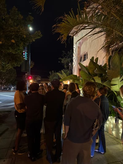 A group of people stand on a sidewalk at night in front of a building. The building appears to be a restaurant or bar, with large tropical plants in front. The street and sidewalk are wet, suggesting it may have recently rained. A traffic light is visible in the background, with a red light illuminated. The people are casually dressed and appear to be waiting in line or socializing. One person in the foreground is looking at their phone. The overall atmosphere is relaxed and social. The location is on South Coast Highway 101 in Encinitas, California, United States of America. The name "SANTA" is visible on the building's facade.