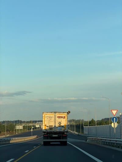 A truck is driving down a highway in Kaipy, Russia. The sky is blue with some clouds. The truck is white and has writing on the back. There are trees and other signs along the side of the road.