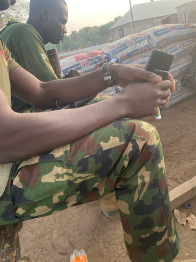 A person wearing camouflage pants and a green shirt is holding a phone in their hands. The person also wears a camo watch on their wrist. They appear to be sitting down. Behind the person are stacked bags labeled "DANCOTE 3X CEMENT". To the side of the person are visible patches of ground and what appears to be a water bottle.