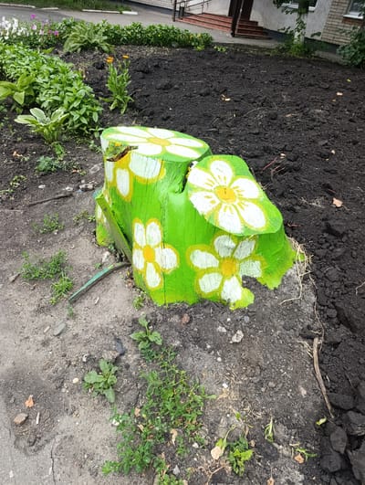 The image shows a green tree stump painted with white and yellow flowers, situated in a garden bed filled with dark soil and some green plants. The stump appears to be in an outdoor setting, likely a park or residential area, given the presence of a building in the background. The building has a red-carpeted staircase leading up to a doorway and is partially visible, suggesting an urban environment. The overall scene depicts a decorative element in a planted area.