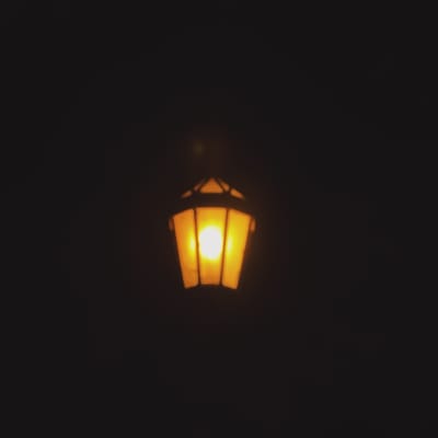 A single illuminated lantern hangs suspended in the dark. The lantern appears to be of a geometric, possibly hexagonal, design with glowing yellowish-orange panels revealing the light source inside. The surrounding environment is entirely dark, with no discernible background details visible. It is impossible to determine if this image was taken in San Fernando del Valle de Catamarca, Argentina, or to provide any further specific details without additional context.