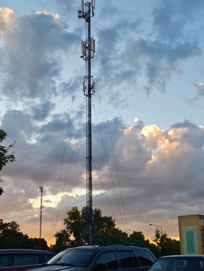 This media file depicts a cell tower against a dramatic sky. The tower is tall and metallic, with several antennas and equipment mounted on it. It is supported by guy wires that extend to the ground. In the foreground, several vehicles are parked, partially obscuring the base of the tower and a line of trees. The sky is filled with a mix of clouds, some dark and grey, others illuminated with the warm hues of sunset. The sun is setting, casting a golden glow on some of the clouds and the upper parts of the tower. The overall atmosphere is serene and slightly dramatic due to the contrast between the industrial structure and the natural beauty of the sky. The location is identified as Svietlahorsk, Belarus. The image captures a moment during sunset, with cloudy weather. There is no visible text in the image.