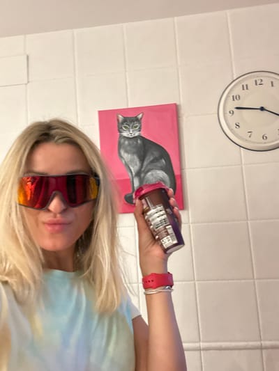 A blonde woman wearing oversized, reflective sunglasses and a tie-dye t-shirt is holding a purple bottle with a red lid. She is standing in front of a tiled wall, to the left of a painting of a grey cat on a pink background. A round clock is visible on the wall to the right of the cat painting. The woman is also wearing a pink smartwatch and a silver bracelet. The scene appears to be indoors, likely in a bathroom or kitchen based on the tilework. There is no indication of the specific location within the city of València, Spain.