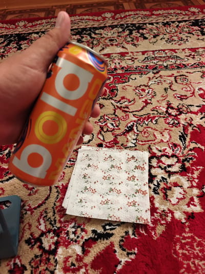 An overhead, medium shot shows a person's thumb and index finger holding an orange soda can. The can is mostly silver with orange lettering that says "BOLA". To the right of the can, a stack of four floral patterned paper napkins are visible. The napkins have a white background with small red and green flowers. All of these items are placed on a red and beige patterned rug. In the lower left corner, a small portion of a blue object is visible. The background of the image is a wooden floor.