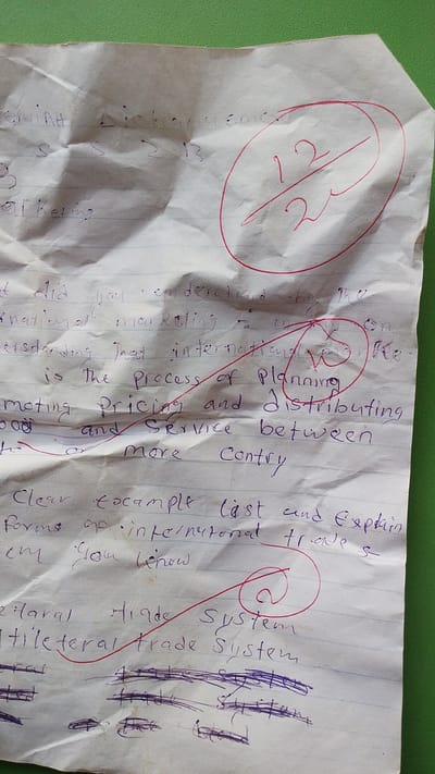 This is a close-up, overhead shot of a crumpled piece of lined paper with handwritten text in blue ink, placed on a green surface. The paper appears to be a school assignment or notes, given the nature of the writing and the presence of a grade circled in red ink.
The text on the paper discusses concepts related to international marketing and trade. Visible phrases include "international marketing," "process of Planning," "pricing and distributing," "Service between," and "Forms of international trade." There is a handwritten grade of "12/22" circled in red ink, suggesting a score or evaluation. Several other parts of the paper have been crossed out or marked with red ink, including a circled "@" symbol and a red line drawn through some of the text.
The handwriting is legible, though somewhat rushed. The crumpled state of the paper and the casual writing style suggest it might be personal notes taken during a lesson or a study session. The green background could be a table, desk, or floor. There are no visible people in the image, and the context suggests an indoor or at least a controlled environment where such notes would be taken. The specific location cues from Donga, Nigeria, are not evident in the visual elements of the image itself.
The overall impression is that of a student’s academic work, possibly a draft or a graded assignment, with an emphasis on business or economics terminology. The red markings indicate areas of focus, correction, or scoring.