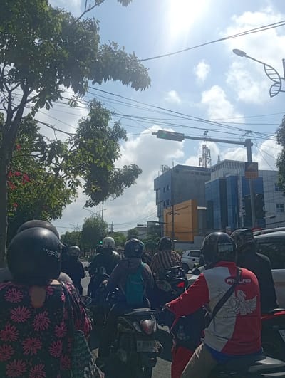 The image captures a street scene in Surabaya, Indonesia, under a bright, sunny sky with scattered clouds. The perspective is from behind a group of motorcyclists stopped at a traffic light. In the foreground, the backs of several individuals wearing helmets are visible. One person on the left wears a black helmet and a black shirt with a vibrant pink floral pattern. To their right, a person in a black helmet wears a striped shirt. Further ahead, other motorcyclists are seen, some with helmets of various colors including white and dark blue.
The environment features lush green trees lining the left side of the street, with some red flowers adding a splash of color. Overhead, a complex network of electrical wires crisscrosses the sky. A traffic light, currently showing green, is mounted on a pole spanning across the road. In the background, modern buildings with blue and grey facades dominate the skyline, one of which displays a CCTV sign. A prominent radio tower is visible on top of one of the buildings. The overall scene depicts a typical urban traffic situation in Surabaya during the daytime.