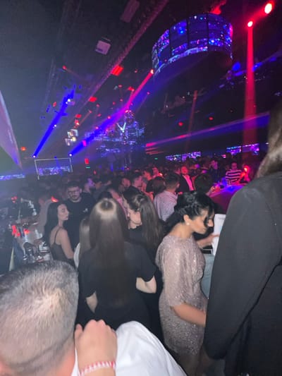 This image captures a densely populated nightclub or party venue located in PAMPOROVO, Bulgaria. The environment is dark, illuminated by a dynamic array of blue and red laser beams, spotlights, and ambient lighting originating from the ceiling. A large, multifaceted disco ball hangs centrally in the upper mid-ground, reflecting light across the scene. A prominent, curved LED screen displaying animated patterns is suspended from the ceiling in the upper right, with a smaller rectangular screen visible in the upper left.
The scene is crowded with numerous individuals, primarily young adults, standing in close proximity to one another. In the immediate foreground, the back of a person's head and right shoulder, clad in a white shirt, are visible, with a red and white striped bracelet on their wrist. In the mid-ground, a woman with long, dark hair, wearing a black top, is observed from behind, facing away from the camera. To her right, a woman with dark hair styled with braids, wearing a shimmering, light-colored (possibly rose gold or silver) dress with short sleeves, is depicted in profile, holding a light-colored object, likely a drink or phone. Another woman with dark hair and a dark top is visible to the left of the woman in black, looking left. Scattered throughout the background, groups of people are visible, some engaged in conversation, with their faces and upper torsos discernible amidst the dim lighting. On the left side of the frame, several small tables with bottles and glasses are partially visible, suggesting seated areas or VIP sections. The overall scene depicts a lively and active social gathering within a club setting.