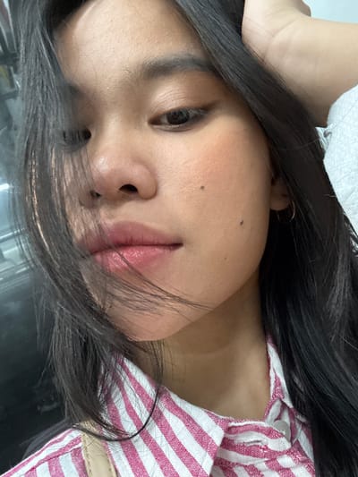 The image is a close-up, vertically oriented selfie of a young woman. She has long, dark hair that falls across her face and shoulders, obscuring parts of the frame. Her head is tilted to the side, and her eyes are looking towards the camera. She is wearing a pink and white striped shirt, and a light-colored fabric is visible in the upper left corner of the image. One of her hands appears to be resting on her head. The woman has on makeup, including what appears to be lip gloss and blush. There are a couple of small moles visible on her face. The lighting suggests it is daytime, possibly indoors, given the quality of the light. There are no visible text elements in the image. The overall impression is a casual, personal snapshot.