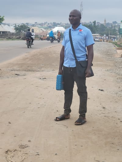 An image captures a full-body shot of a man standing on an unpaved road in what appears to be a semi-urban or rural setting in Suleja, Nigeria. The man, with a bald head and a neutral expression, looks directly at the viewer. He wears a light blue polo shirt featuring the "Pepsi" logo on the left chest, dark trousers, and dark sandals. A black shoulder bag crosses his torso, and he holds a small, rectangular blue object in his left hand, down by his side.
The road he stands on is sandy/dusty with scattered small stones, and it appears to be a shoulder or side lane next to a slightly more defined but still unpaved main road. In the background, there is a busy scene with multiple motorcycles, some carrying passengers, and what looks like a few cars. Buildings, ranging from single-story structures to more substantial ones, are visible along the roadside and stretching into the distance. A tall, light-colored structure, possibly a mosque or tower, is visible among the buildings. The sky is overcast or hazy, suggesting a bright but not sunny daytime. The overall impression is one of everyday life in a bustling local environment.