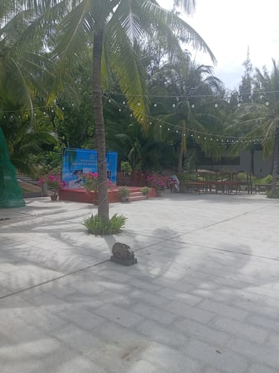 This image captures an outdoor scene at what appears to be a resort or event space in Dong Ninh Hoa, Vietnam. The foreground is dominated by a large, paved courtyard made of light gray rectangular tiles, with shadows of palm trees cast across it. To the left, a green mesh structure is visible.
In the mid-ground, a raised red stage is set up with a blue backdrop displaying text in Vietnamese, including "CHÀO MỪNG QUÝ KHÁCH" and "GM DOC LET BEACH RESORT & SPA". Two potted plants with vibrant pink flowers flank the stage. A single palm tree with a textured trunk, wrapped with white rope, stands prominently in front of the stage. Behind the stage, stacks of red chairs are visible, and further back, several wooden tables and chairs are arranged.
The background consists of dense tropical foliage, primarily palm trees and other greenery, suggesting a warm climate. String lights with white bulbs are hung across the trees, indicating preparation for an evening event or ambiance. The sky is overcast, with a bright, diffused light. A person in a white outfit with a red sash is partially visible near the tables and chairs in the background, possibly an employee or attendee. In the far right, there are more stacks of red chairs. On the tiled ground in the lower right quadrant, a rusted metal cutting tool sits on a dark base.
